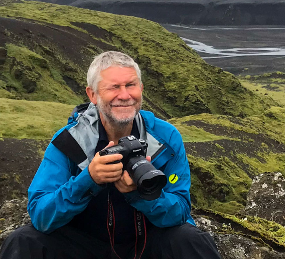Foto på Jim Sundberg som sitter på huk med en kamera i händerna i ett grönt landskap.